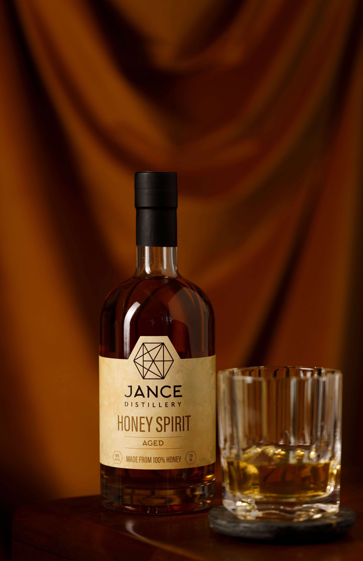 Coupla Jance Distillery aged honey spirit in a glass bottle next to a crystal tumbler of amber spirit on a coaster.
